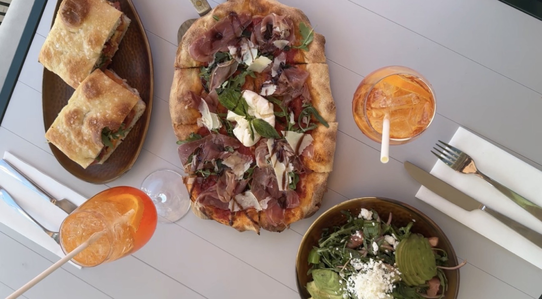 Overhead patio table with prosciutto pizza, focaccia sandwich, Aperol spritz and an avocado salad